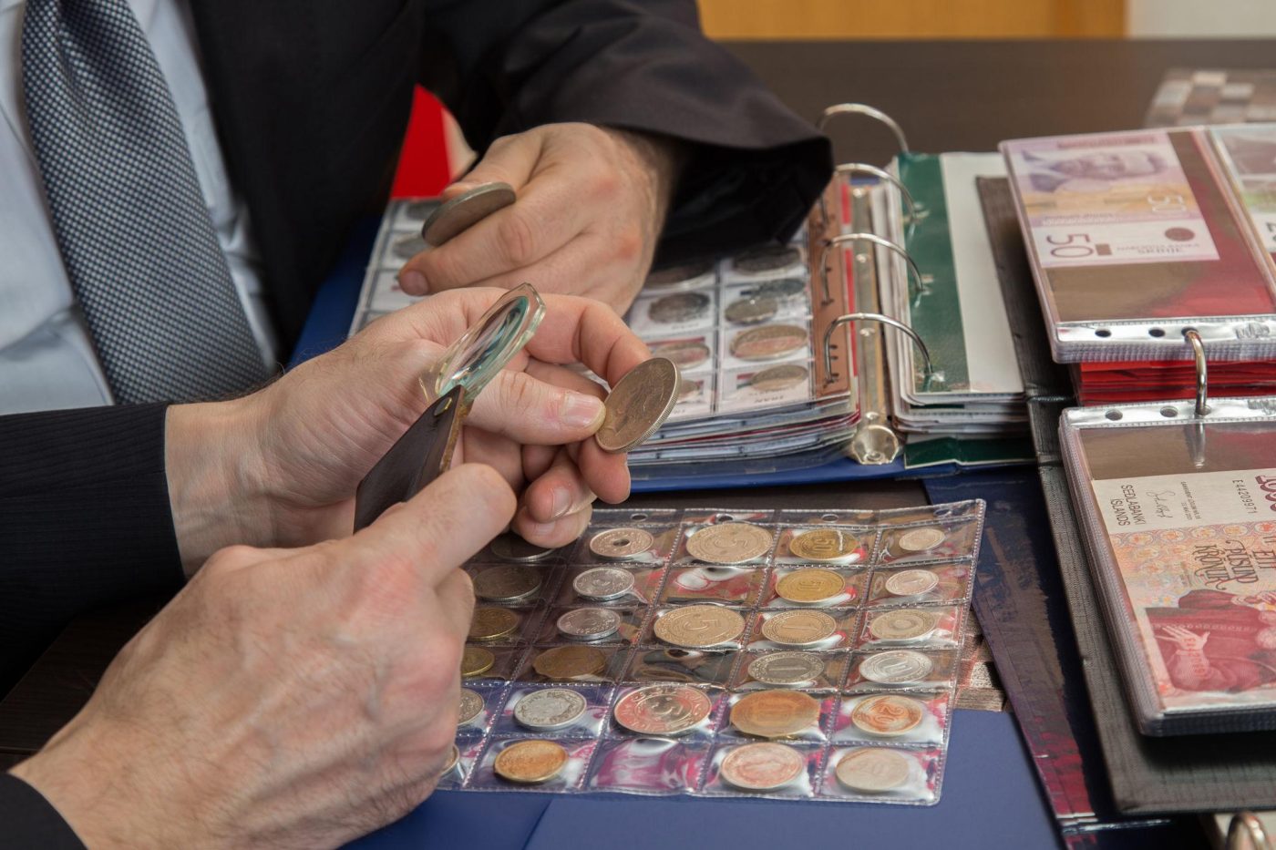 Two collectors reviewing coins with a magnifying glass next to albums and currency